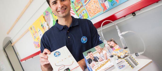 Ein Mann in einem blauen Polohemd hält ein TUEV Kids Strombausets "TUEV Kids", die ein Spielzeugauto und eine Bedienungsanleitung enthält. Hinter ihm sind bunte Kunstwerke an der Wand zu sehen.