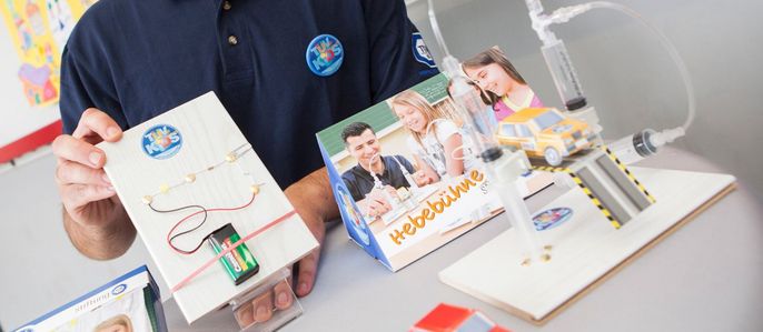 Ein Mann in einem blauen Polohemd hält ein TUEV Kids Strombausets "TUEV Kids", die ein Spielzeugauto und eine Bedienungsanleitung enthält. Hinter ihm sind bunte Kunstwerke an der Wand zu sehen.