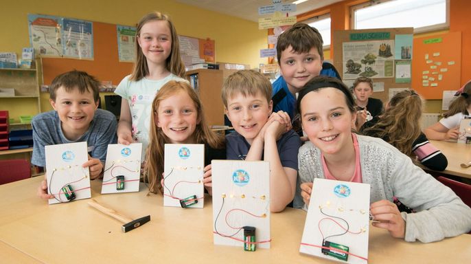 Eine Gruppe von Kindern in einem Klassenzimmer, die Karten mit dem TUEV Kids Logo und einem Schaltkreis hochhalten, mit einem Holzhammer auf dem Tisch.