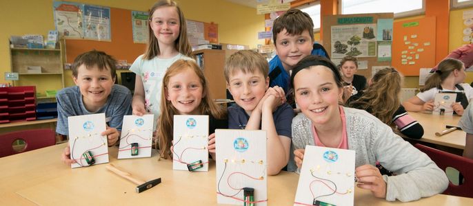 Eine Gruppe von Kindern in einem Klassenzimmer, die Karten mit dem TUEV Kids Logo und einem Schaltkreis hochhalten, mit einem Holzhammer auf dem Tisch.