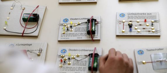 Verschiedene elektronische Schaltkreise, die jeweils mit TUEV Kids Stickern und einer kurzen Beschreibung versehen sind, zusammen mit einer Batterie und Anschlusskabeln.