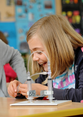 Ein junges Mädchen mit blondem Haar, das lächelt und auf eine TUEV Kids Hebebühne auf einem Tisch schaut.