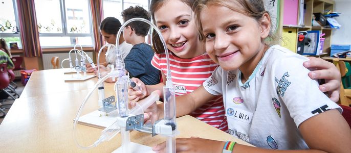 Zwei junge Mädchen in einem Klassenzimmer, lächelnd und in die Kamera blickend, vor denen eine TUEV Kids Hebebühne aufgebaut ist.