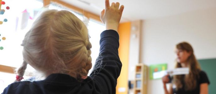Ein junges Mädchen mit blondem Haar, das in einem Klassenzimmer die sich meldet, während eine Frau im Hintergrund Unterricht hält.