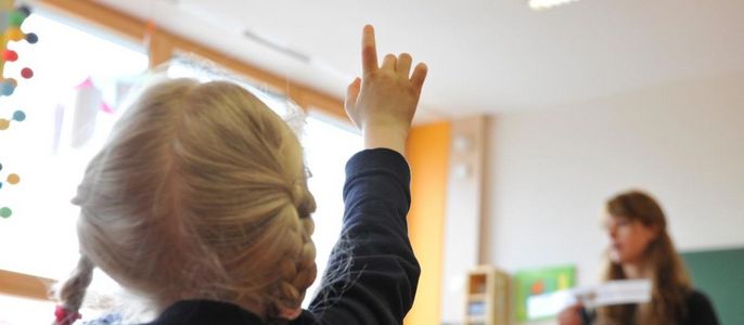 Ein junges Mädchen mit blondem Haar, das in einem Klassenzimmer die sich meldet, während eine Frau im Hintergrund Unterricht hält.