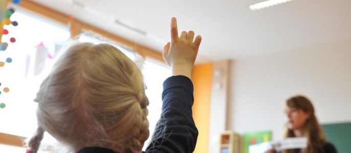 Ein junges Mädchen mit blondem Haar, das in einem Klassenzimmer die sich meldet, während eine Frau im Hintergrund Unterricht hält.