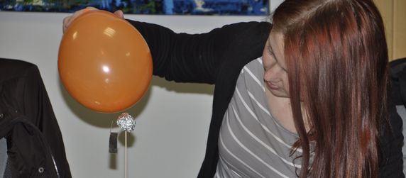 Eine Frau mit langen roten Haaren, die einen orangefarbenen Luftballon über einem Tisch mit verschiedenen Gegenständen, darunter Drähte und eine Batterie, hält.