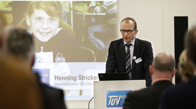 Henning Stricker im Anzug steht auf einem Podium und präsentiert vor einem Publikum, auf einem großen Bildschirm ist ein junges Mädchen zu sehen.