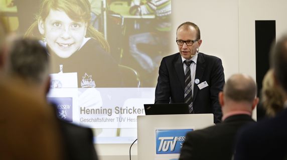 Henning Stricker im Anzug steht auf einem Podium und präsentiert vor einem Publikum, auf einem großen Bildschirm ist ein junges Mädchen zu sehen.