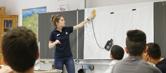 Eine Frau in blauem Hemd und Jeans steht vor einer Tafel und erklärt einer Gruppe von Jungen, die an Tischen sitzen, etwas.