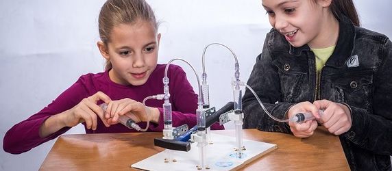 Zwei junge Mädchen arbeiten an einer TUEV Kids Hebebühne, wobei ein Mädchen ein Werkzeug hält und das andere beobachtet.