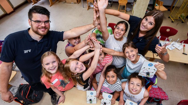 Eine Gruppe von Kindern und Erwachsenen in einem Klassenzimmer, die alle lächeln und ihre Hände gemeinsam in einer fröhlichen Geste heben.
