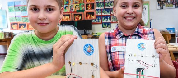 Zwei kleine Kinder in einem Klassenzimmer, jedes hält ein weißes Holzbrett mit einem Schaltkreis, der an eine Batterie angeschlossen ist.
