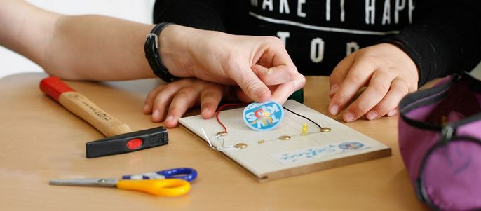 Die Hände einer Person arbeiten an einem Bastelprojekt, auf dem Tisch liegen ein Abzeichen, eine Schere und ein Hammer.