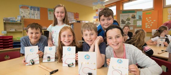 Eine Gruppe von Kindern in einem Klassenzimmer, die Karten mit einem TUEV Kids Motiv hochhalten, umgeben von Unterrichtsmaterialien und Werkzeugen.