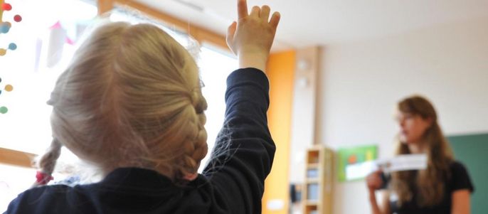 Ein junges Mädchen mit blondem Haar, das in einem Klassenzimmer die Hand hebt, während eine Frau in der Nähe steht und ein Stück Papier hält.