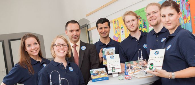 Eine Gruppe von sieben Personen, einige in blauen Polohemden mit dem Logo "TUEV Hessen", steht um einen Tisch herum, auf dem verschiedene Produkte ausgestellt sind, darunter ein TUEV Kids Strom Bauset.
