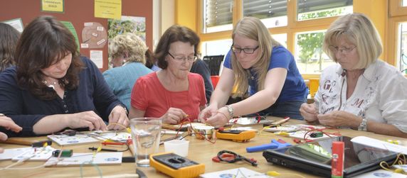 Vier Frauen arbeiten an elektronischen Projekten auf einem Tisch, umgeben von verschiedenen Werkzeugen und Bauteilen.